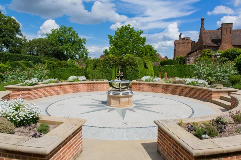 A large sundial set in the centre of a paved area surrounded by a low brick wall, with topiary, trees and a red brick historic building in the background. 