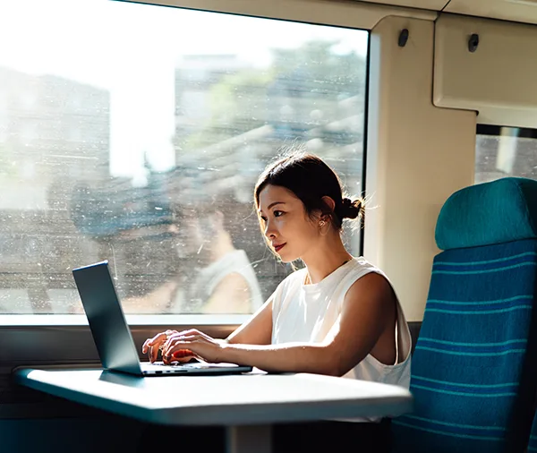 A young East Asian woman on a train, wearing a short sleeved white top and sitting working on a laptop at a table.