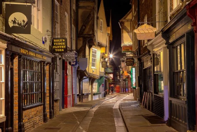 The quaint streets of York's thoroughfare are lit dimly in the evening, with the windows of York's shopfronts framing the image.