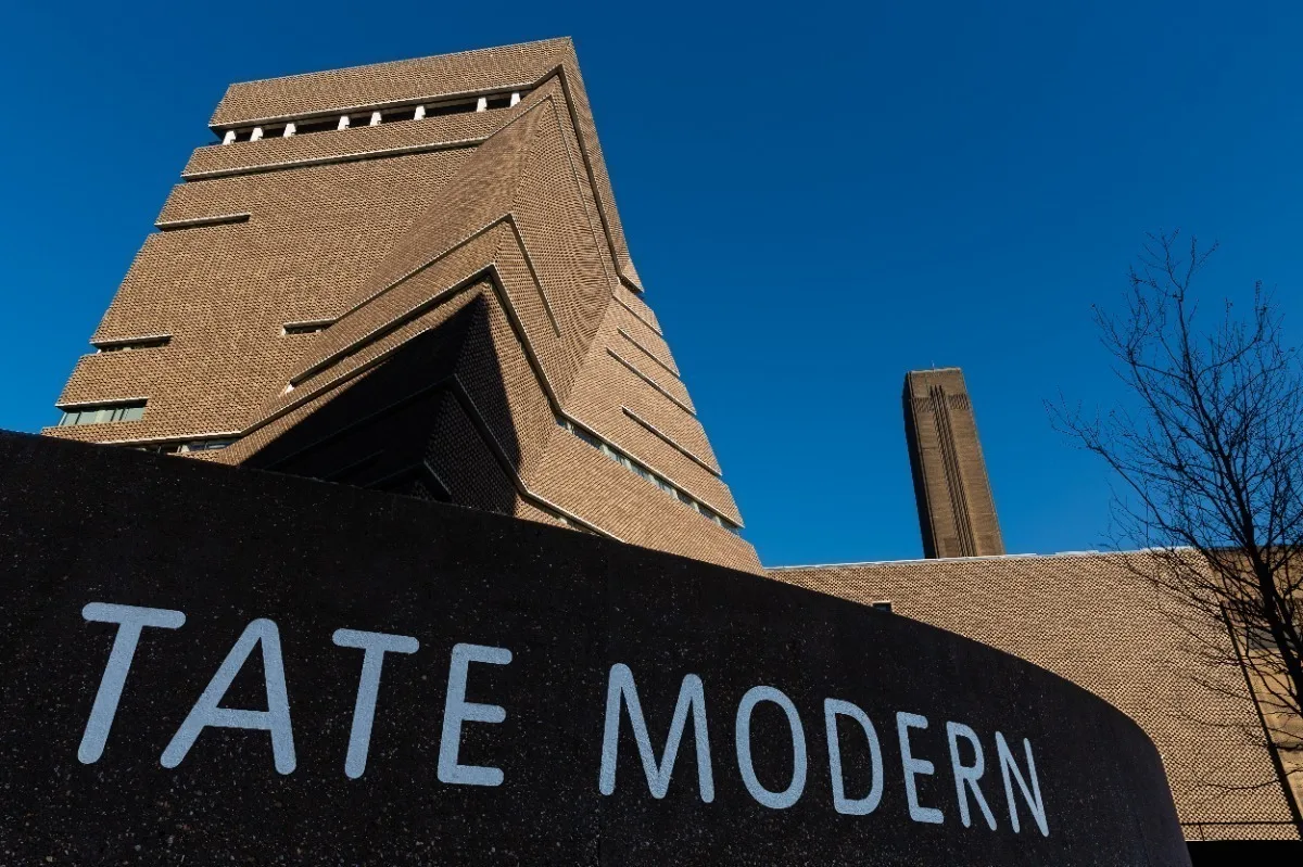 Tate Modern museum's brick tower and chimney against a clear blue sky, with sign with white letters visible in foreground.