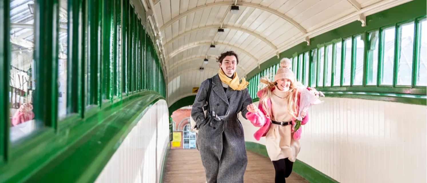 A man and a woman dressed in coats and scarves are holding hands and laughing as they run down an enclosed railway bridge with windows either side.