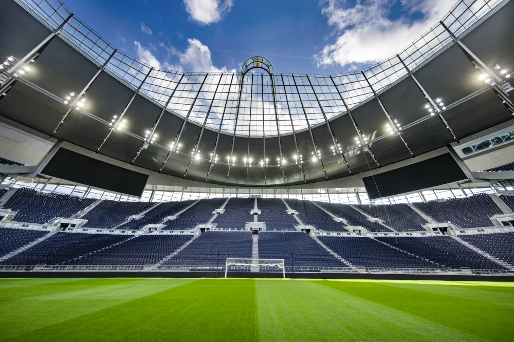 Image of the stand of empty seats behind a goal at Tottenham hotspur stadium. The lights of the stadium are lit up brightly shining on the well manicured pitch.