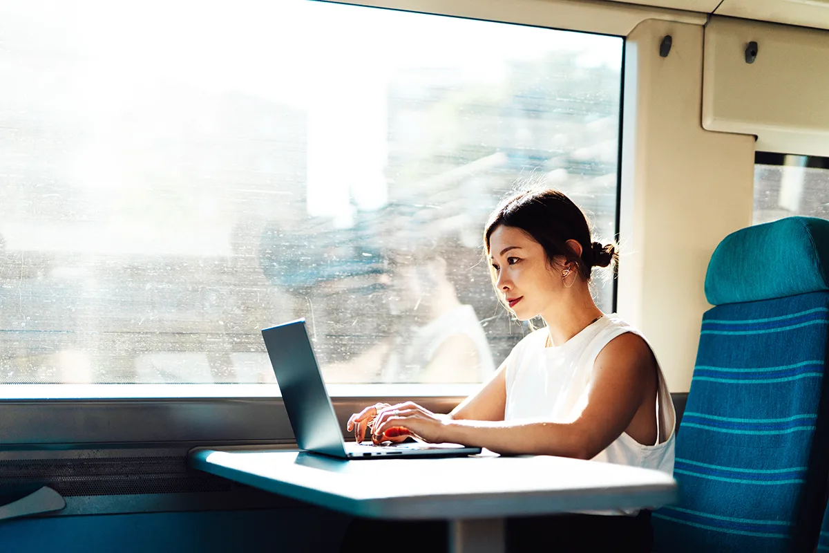 A young East Asian woman on a train, wearing a short sleeved white top and sitting working on a laptop at a table.