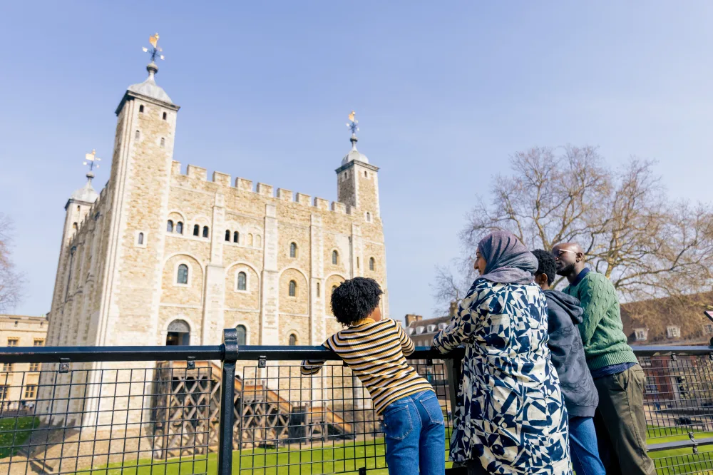 A Black family of 4, leaning on railings and looking at the turreted building of the Tower of London.