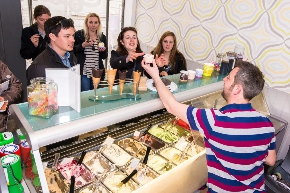 A white man with a beard serving customers behind an ice cream counter in a shop. 