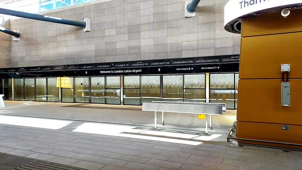 London Luton Airport entrance with glass doors, welcome sign, metal bench, and orange structure to the right.