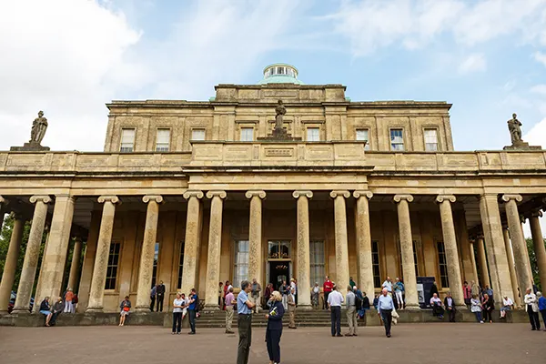 A large yellow stone building with a row of columns across the front and statues on either side of the roof, with people milling around in front. 