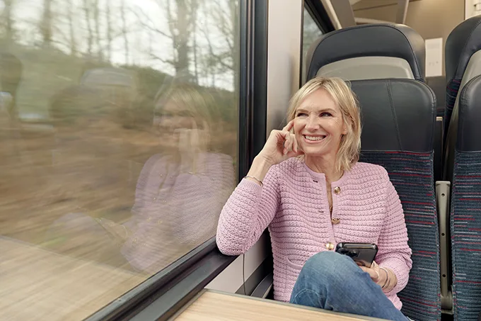 DJ Jo Whiley, a white woman with blonde hair, sitting at a train table holding a mobile phone and smiling as she looks out the window.