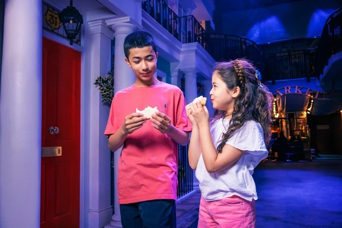 An East Asian boy and girl eating standing and eating sandwiches in an exhibition made to look like a London street. 