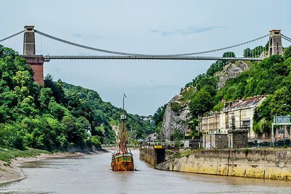 A boat sailing underneath a very high suspension bridge, with green trees and hill either side. 