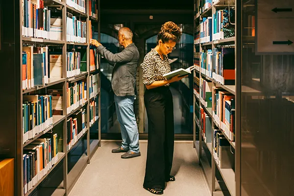 A man and a woman stand in a library looking at books on the shelves.