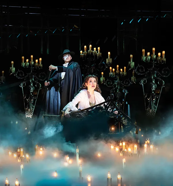 A white woman with long brown hair wearing a flowing white dress sits in a boat being pushed along by a man in a white mask and black hat and cape. They are surrounded by dozens of candles. 