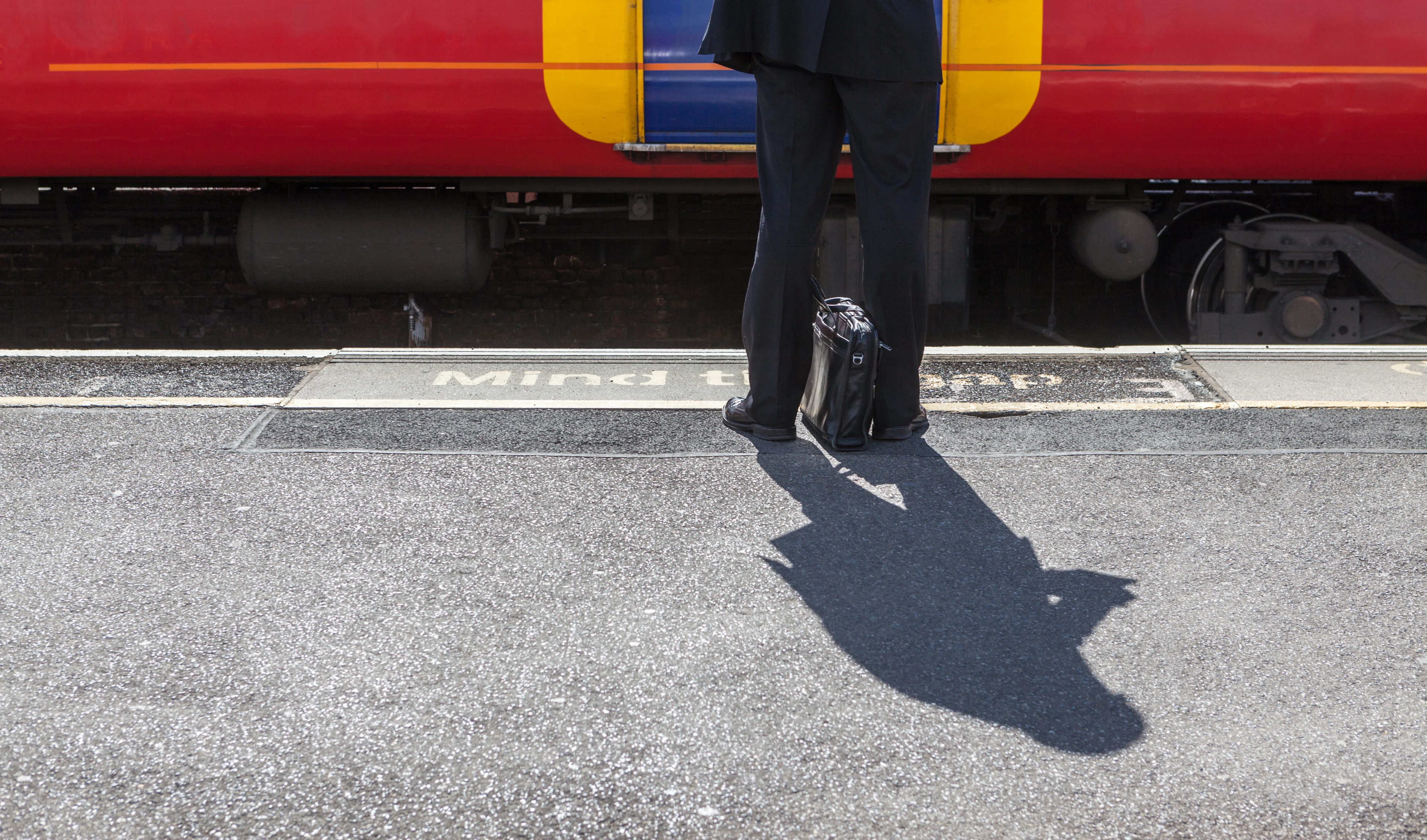 A man with a briefcase on the ground between his legs stands behind the yellow line on a station platform by the Mind The Gap signage on the floor