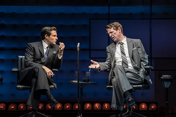 2 white actors in grey suits sitting onstage in a talk-show setting.