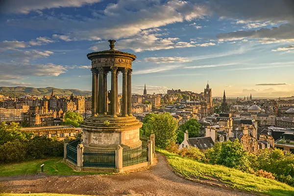 Looking down from a hilltop over the city of Edinburgh, with a stone monument in the foreground. 