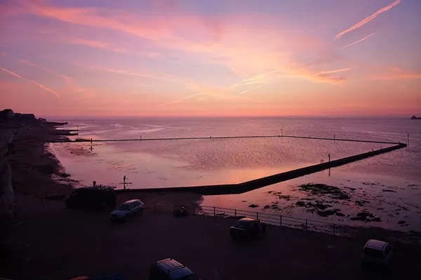 A vibrant pink and purple sunset over a coastal tidal pool, with a sandy beach, calm sea, and parked cars in the foreground.