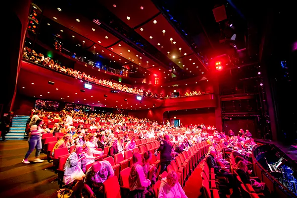 A busy theatre auditorium bathed in red light with audience members filling most of the red velvet seats