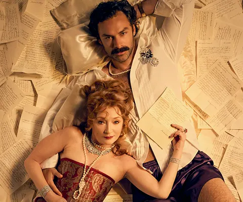 Two people in period costume lying on a bed covered with manuscript pages.