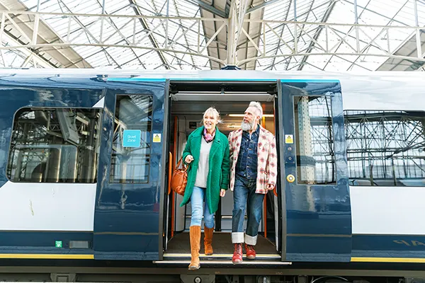 A smiling older couple stepping off a blue train at a station, woman in green coat with brown bag, man in plaid shirt.
