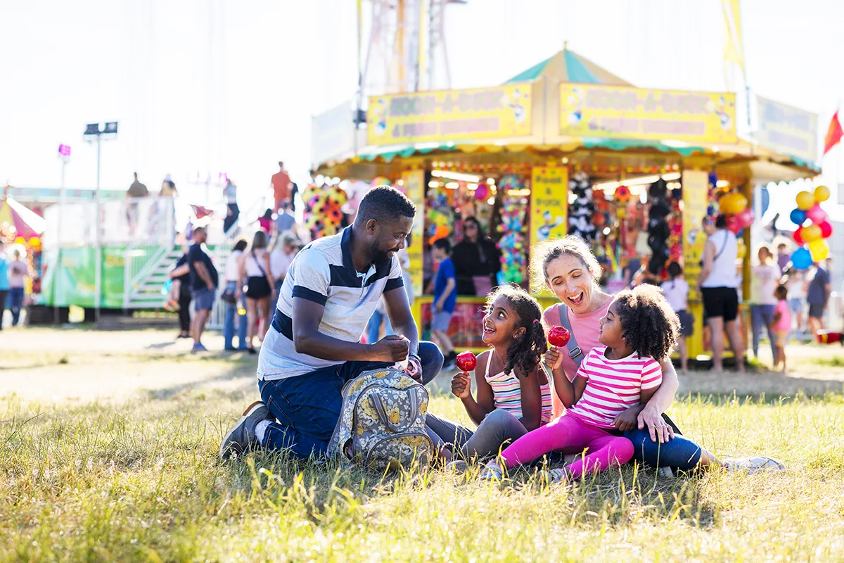 A Black father and white mother with 2 young daughters, laughing and smiling while sitting on grass at a funfair on a sunny day. 