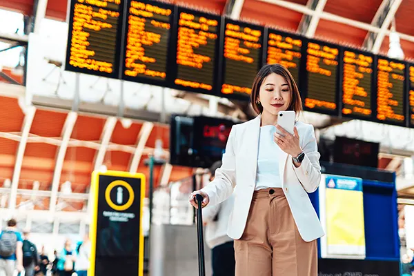 An East Asian woman standing in front of a large departure board at a rail station, looking at her phone and holding a wheeled suitcase.