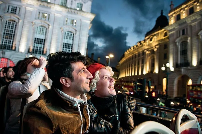 A white couple on the top deck of an open top bus at night, wrapped up in winter clothes, looking at the lights of central London.