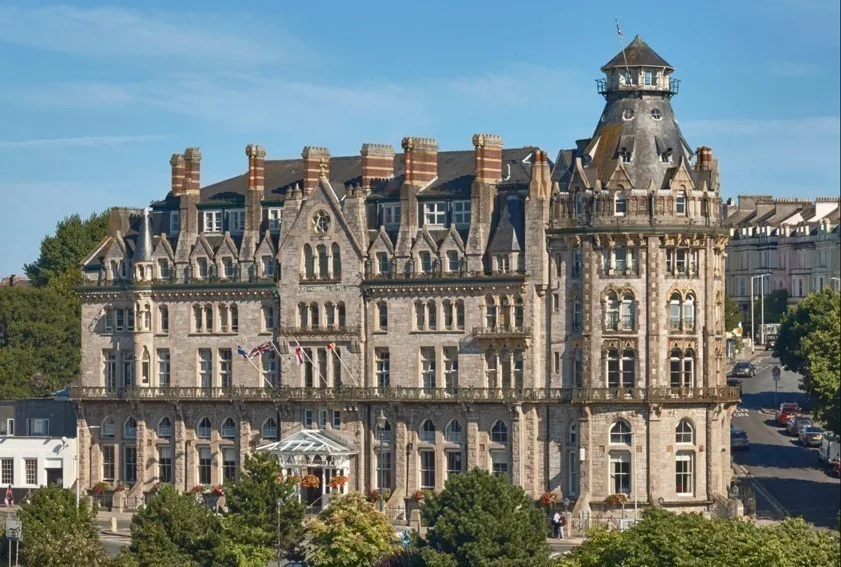 Historic stone hotel with ornate Victorian architecture featuring distinctive turrets and chimneys against a blue sky.