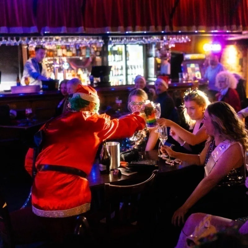 Inside a cosy cabaret bar, a table of women are drinking with a man dressed in a Santa suit. 