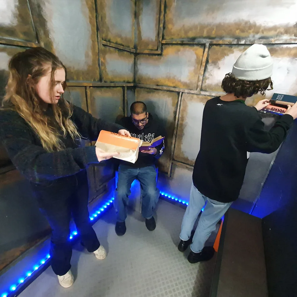 Three people in a dimly lit escape room with metal walls and blue LED floor lighting.