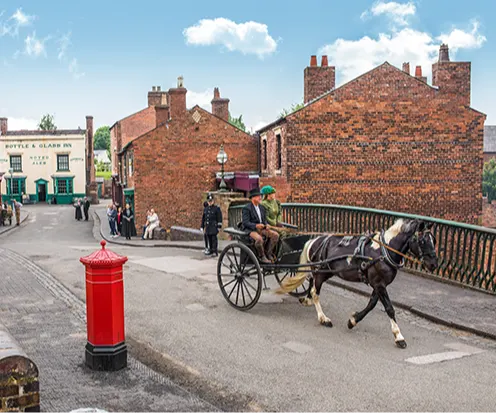 A man and a woman ride in a horse-drawn buggy down a recreation of an old-fashioned street on a sunny day.