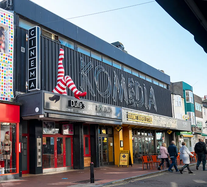 The front of the Komedia arts venue in Brighton, a black-fronted building with an awning that has a large sculpture of red-and-white striped legs attached.