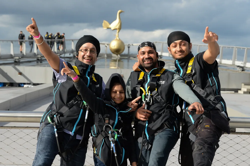4 South Asian people facing the camera and pointing, they are standing on the roof of a football stadium and wearing harnesses and ropes. 