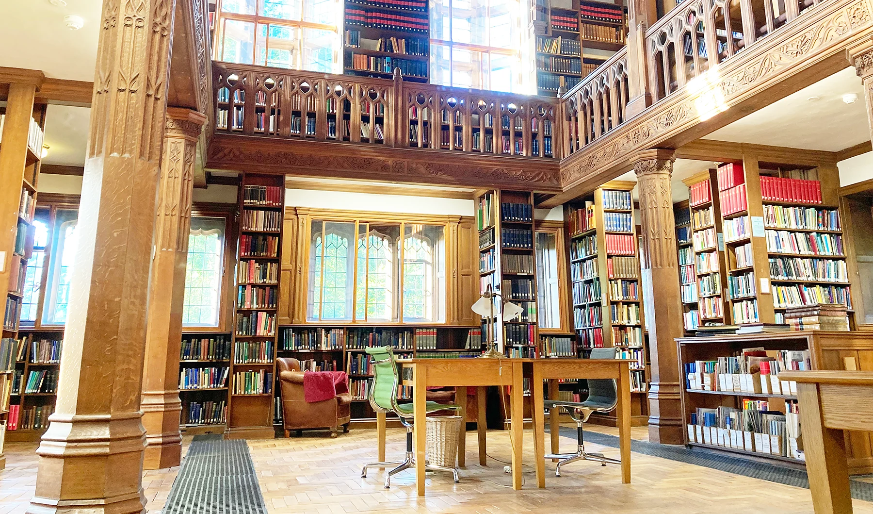 Inside a double-height historical library room with large windows and wooden balcony overlooking a reading area surrounded by bookshelves.