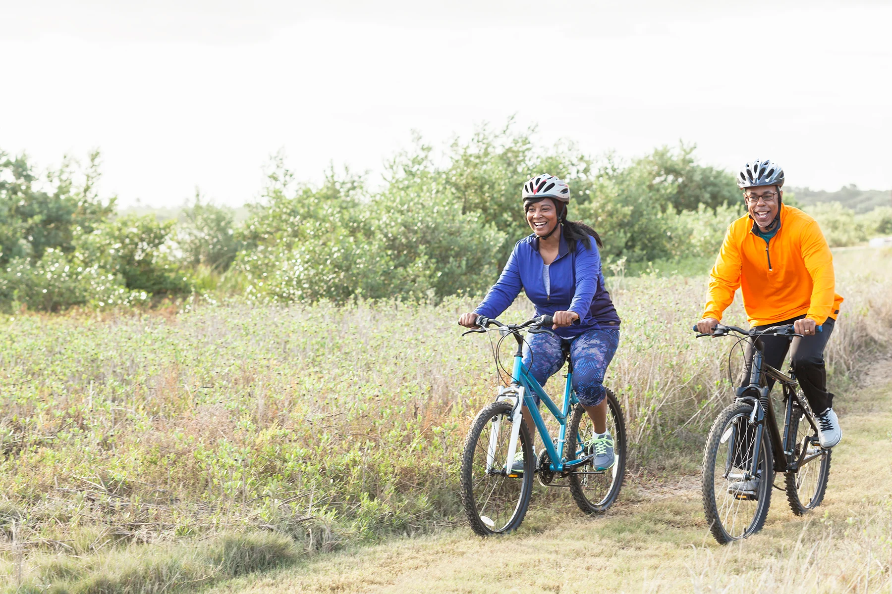 A smiling middle-aged Black woman and Black man in athletic clothing and cycle helmets riding bicycles along a grassy path in the countryside.