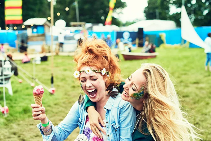 2 white young women sitting on the grass at a festival, they are laughing and one is holding an ice cream cone
