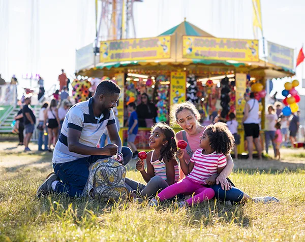 A Black father and white mother with 2 young daughters, laughing and smiling while sitting on grass at a funfair on a sunny day. 