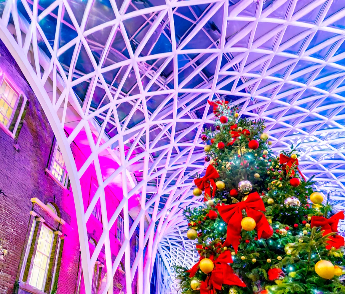 Christmas tree with red bows and gold baubles under a modern geometric ceiling illuminated with purple and blue lights.