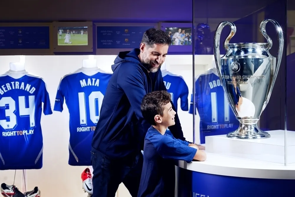 A white man with a beard and a white boy with dark hair looking at a large silver football trophy in a glass cabinet.
