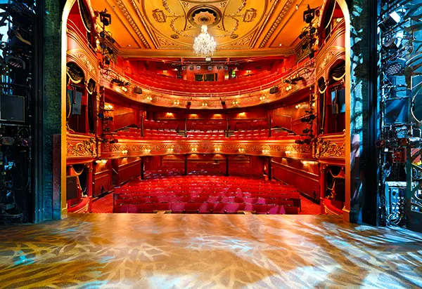 The view from a theatre stage looking into an empty ornate auditorium with rows of red seats.