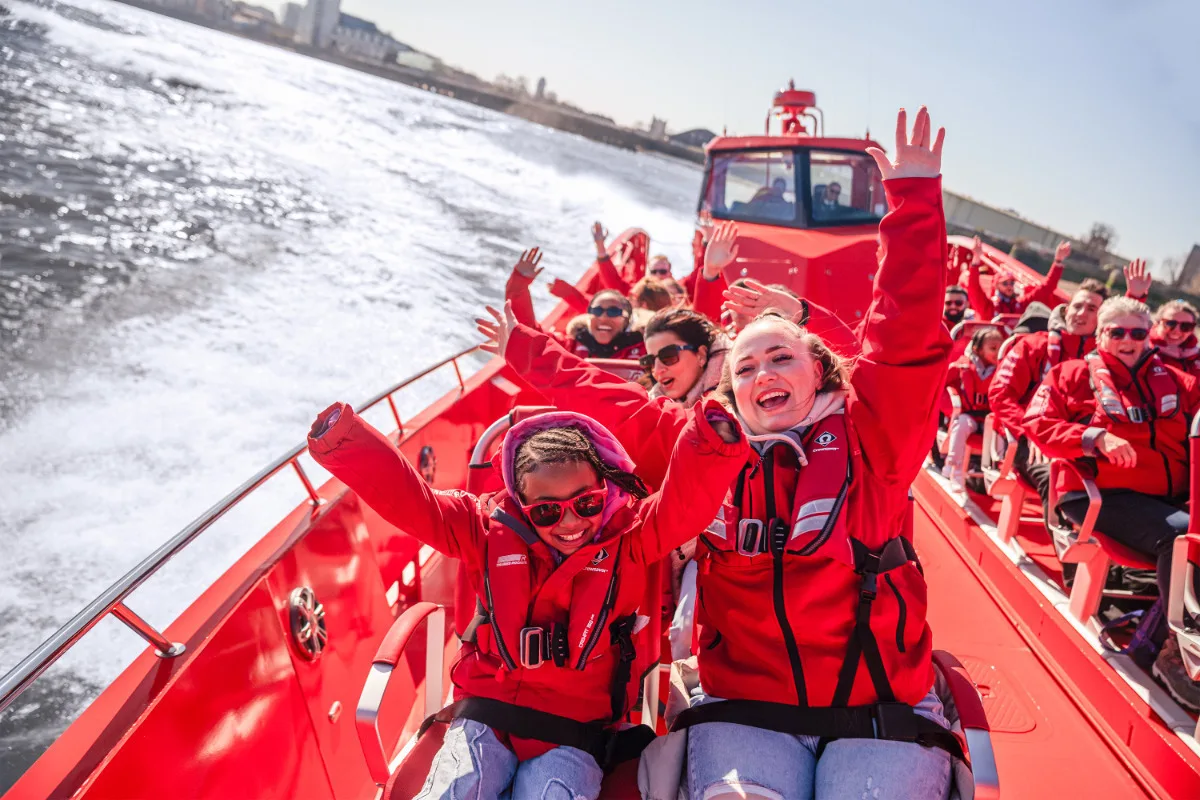 Excited passengers in red life jackets cheer with hands raised on a speeding red boat on the River Thames. 